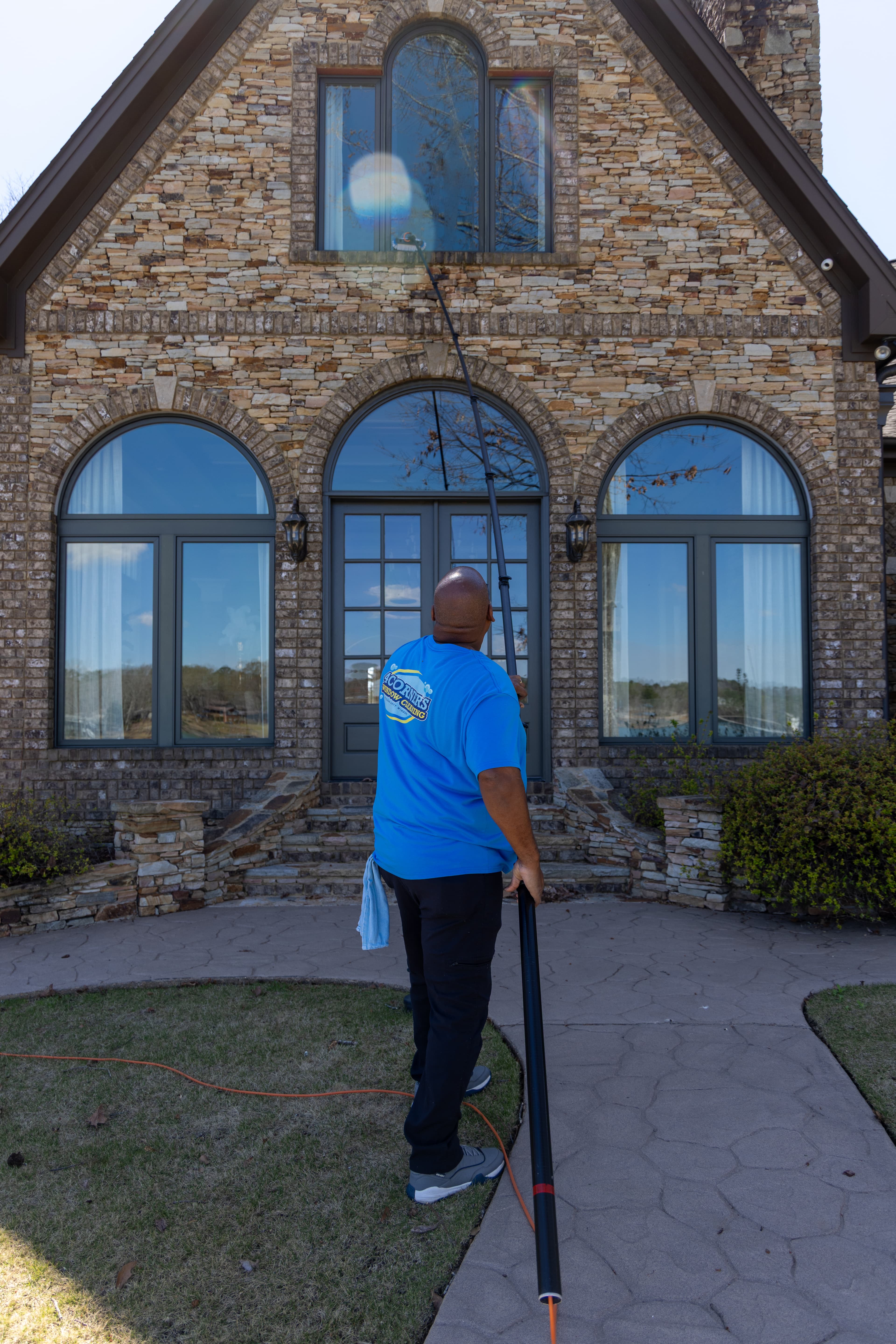 Professional window cleaner using a long pole to wash high windows on a stone house.